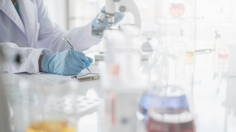 A scientist hands writing on a clipboard in laboratory with test tube microscope and solutions.